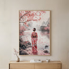 A framed art print depicting a woman in a pink floral kimono standing by a river under blooming cherry blossom trees, displayed on a beige wall above a light wood console table with a small vase and books.
