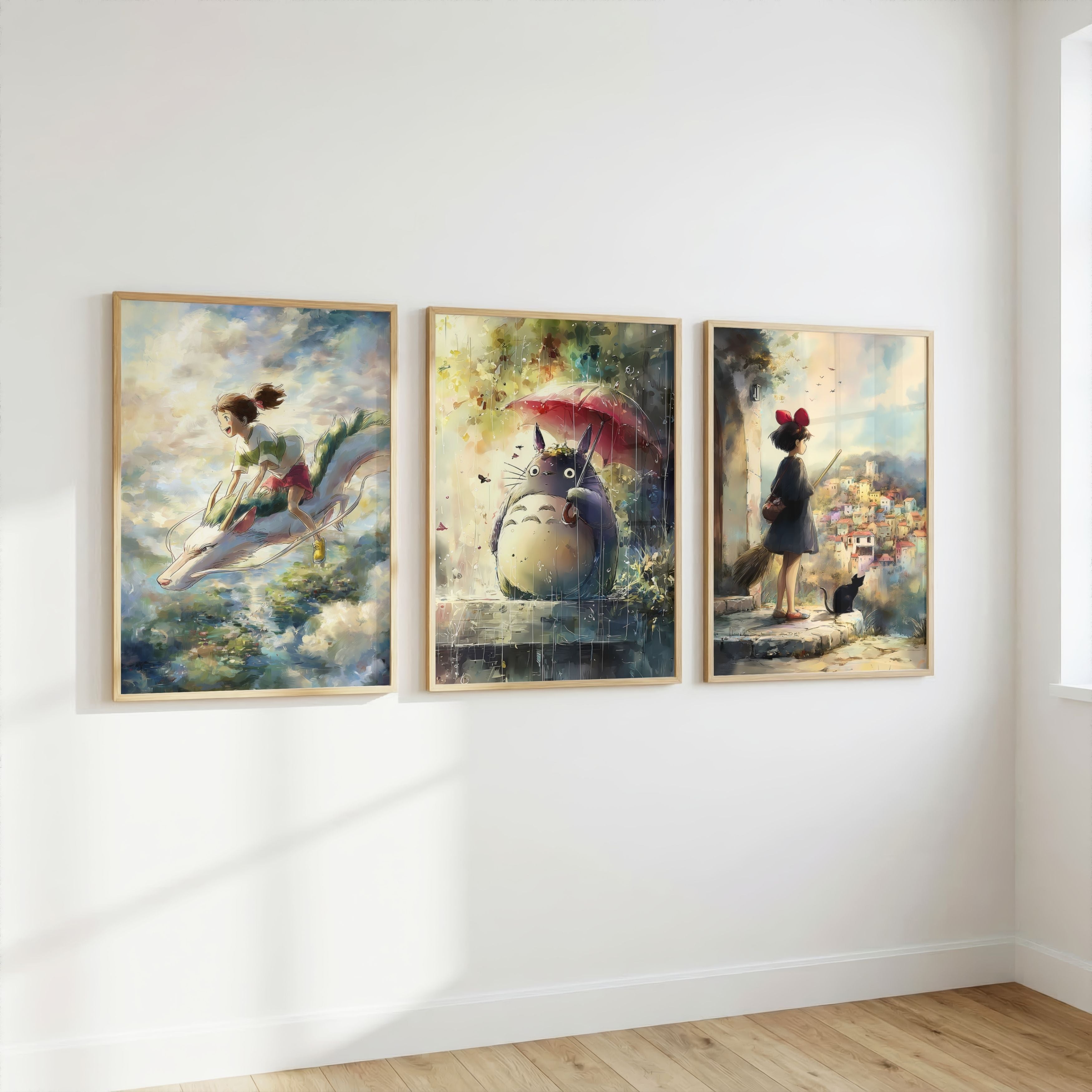 Three framed Spirited Away, My Neighbor Totoro, and Kiki’s Delivery Service in a fantasy paintings hang side by side on a white wall in a room with light wood flooring. The left painting shows a girl riding a dolphin, the center shows Totoro under a red umbrella, and the right shows a girl standing in a garden with a black cat.