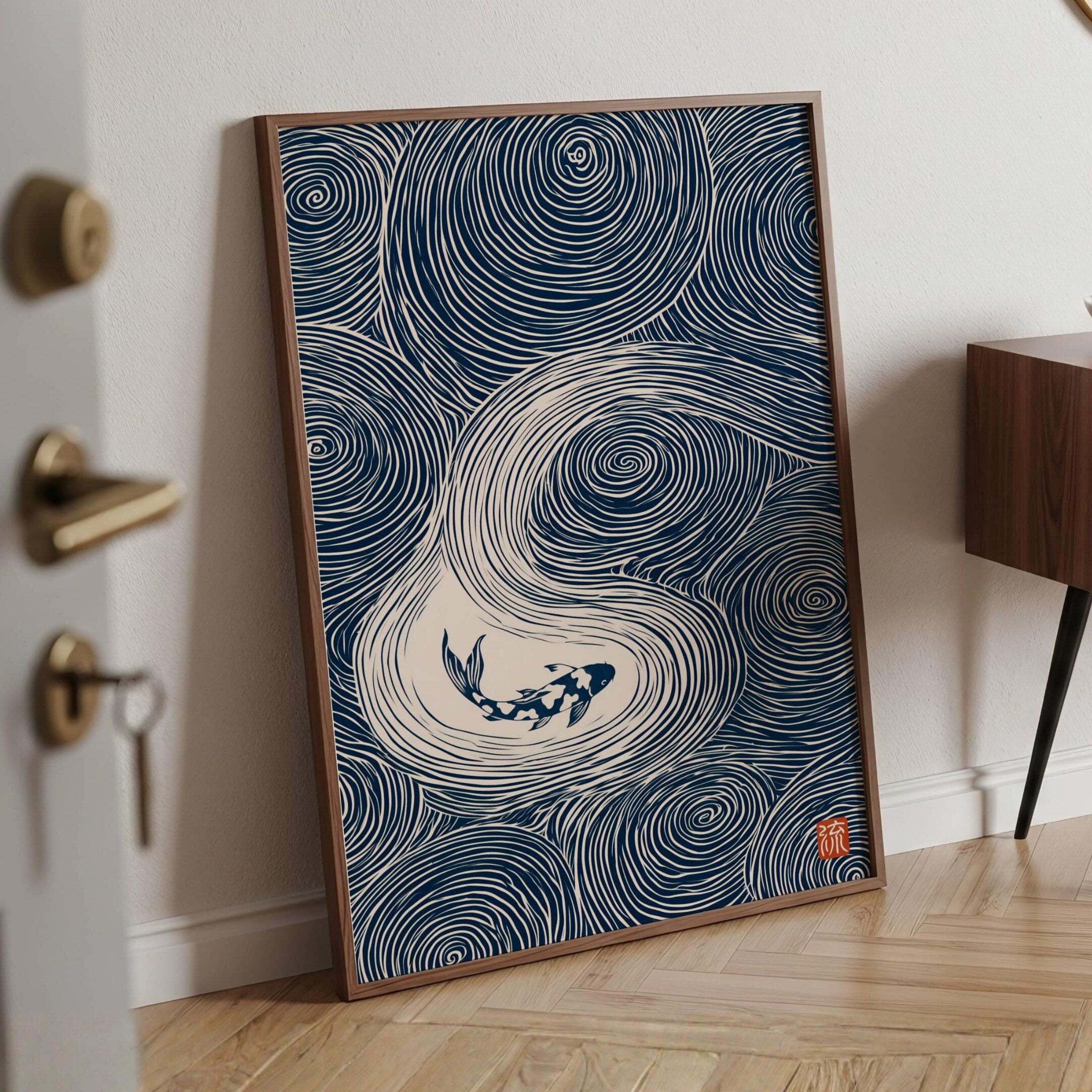 A framed art print featuring a navy blue and white wave design with a koi fish, leaning against a white wall on a light wood herringbone floor.