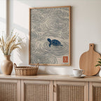 A framed wall art print featuring a blue turtle surrounded by swirling concentric lines on a cream background, styled in a warm, modern interior above a light wood cabinet with woven doors.