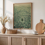 A framed art print featuring a green and beige ripple pattern with a central dark shape, hanging on a light beige wall above a wooden cabinet styled with a vase, basket, cutting board, and cup.