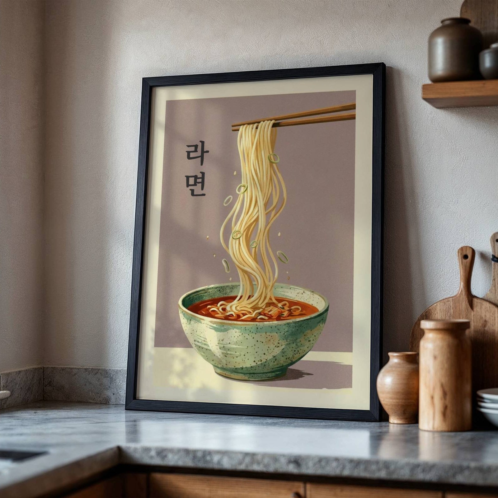 A framed art print featuring a retro-style illustration of Korean ramen noodles hanging from chopsticks above a bowl of red soup, leaning against a kitchen wall on a countertop.