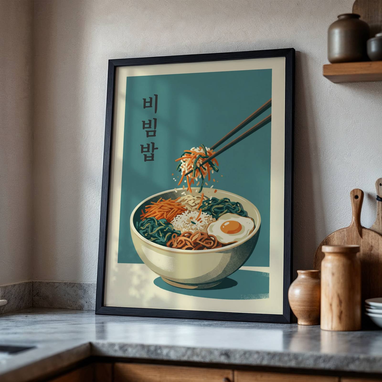 A framed vertical art print of a Korean bibimbap noodle bowl with chopsticks, leaning against a kitchen wall on a countertop.
