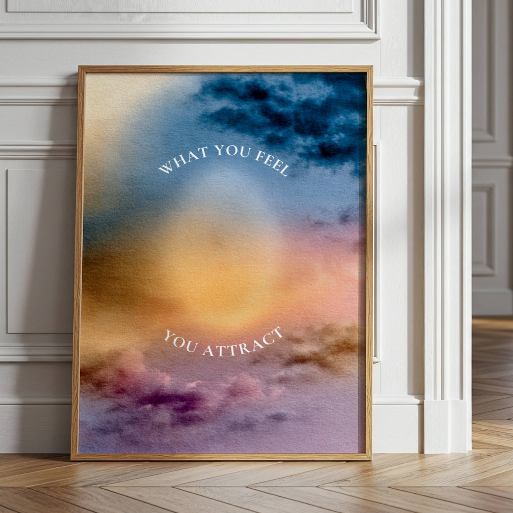 A framed art print leaning against a white paneled wall in a bright, modern room. The print features an abstract watercolor design with a glowing yellow-orange center, blue and purple clouds, and the text 'WHAT YOU FEEL YOU ATTRACT'.