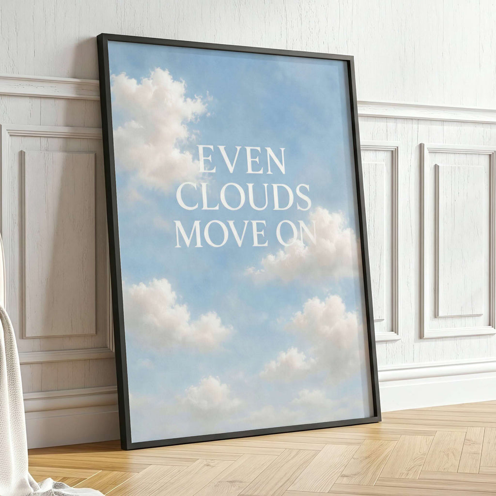 A framed art print leaning against a white paneled wall on a light wood floor, featuring a blue sky with white clouds and the text 'EVEN CLOUDS MOVE ON' in white. A potted green plant stands to the right.