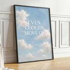 A framed art print leaning against a white paneled wall on a light wood floor, featuring a blue sky with white clouds and the text 'EVEN CLOUDS MOVE ON' in white. A potted green plant stands to the right.