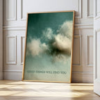 A framed art print leaning against a white paneled wall on a light wood floor. The print features a dark teal sky with soft white clouds and the text 'GOOD THINGS WILL FIND YOU' at the bottom.