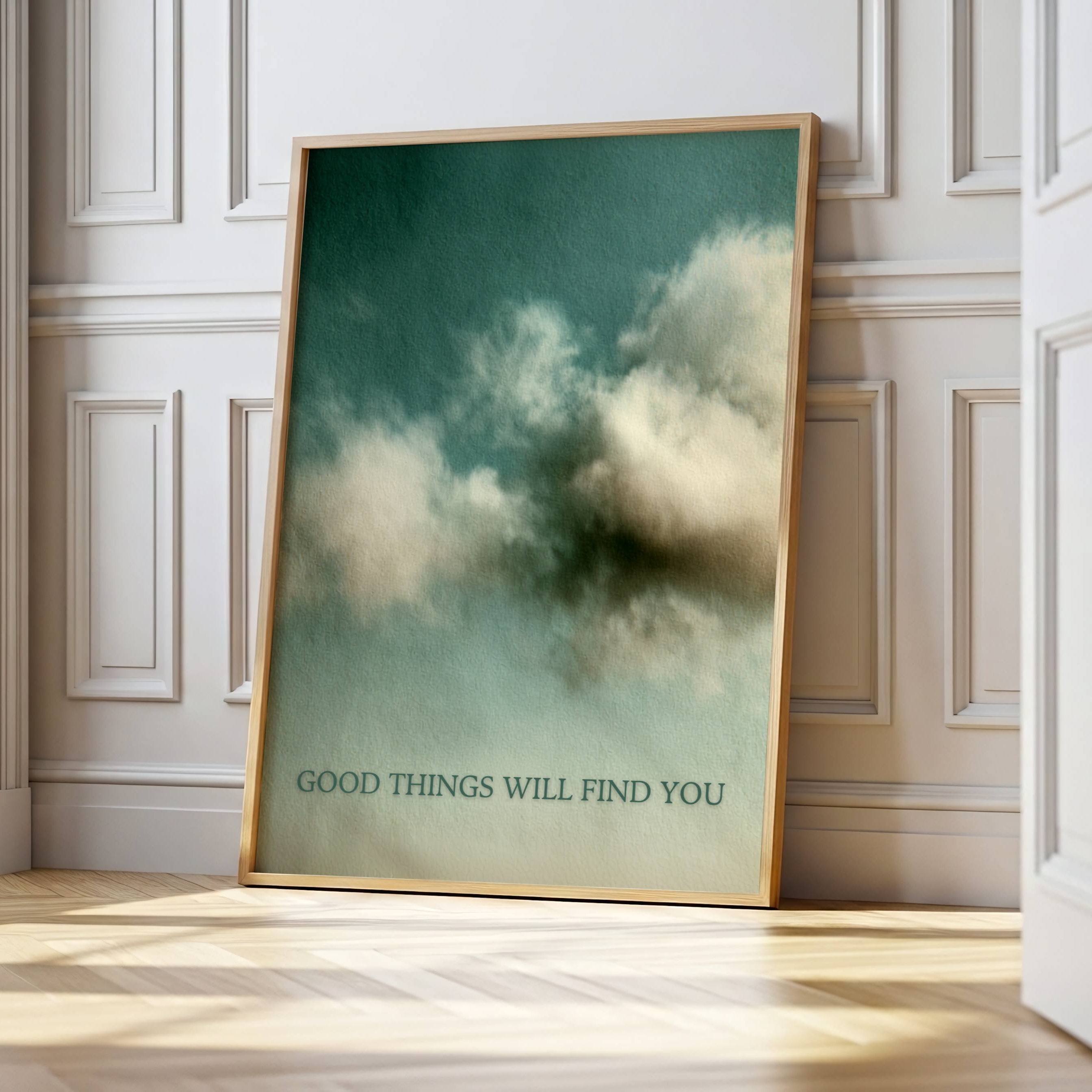A framed art print leaning against a white paneled wall on a light wood floor. The print features a dark teal sky with soft white clouds and the text 'GOOD THINGS WILL FIND YOU' at the bottom.